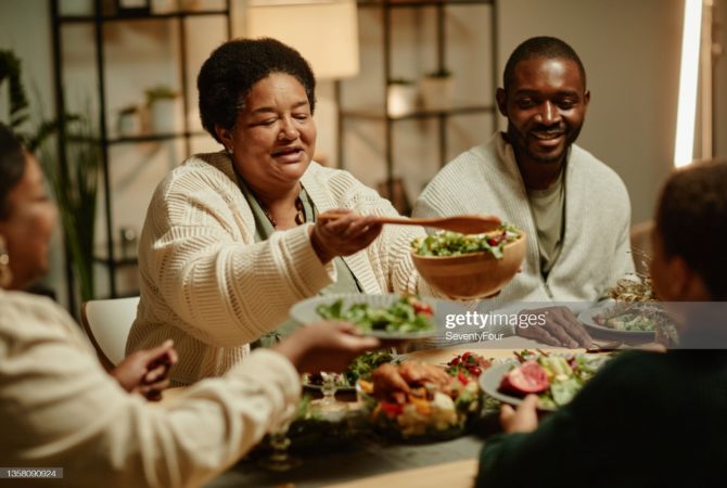 Portrait of smiling African-American grandmother serving food while celebrating Thanksgiving with big happy family at dinner table